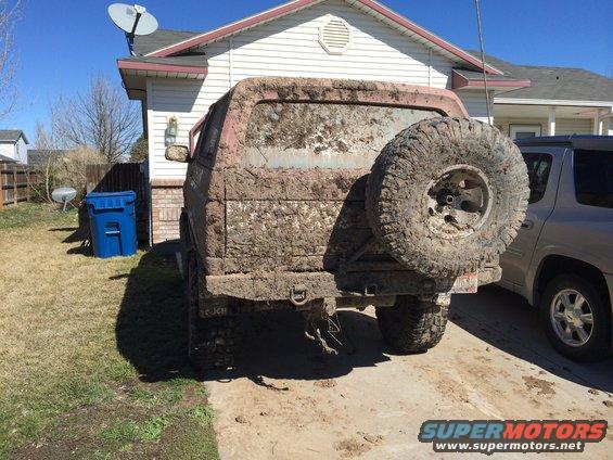 Mud-covered SUV's rear, showing spare tire, tow hitch, and a silver SUV parked nearby