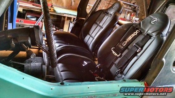 Interior of a teal vehicle with black racing seats and Corbeau harnesses, roll cage visible