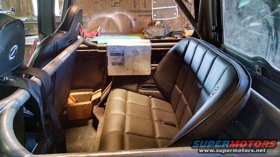 Interior view of a vehicle with aftermarket racing seats and a roll cage. A box sits on a shelf behind the seats