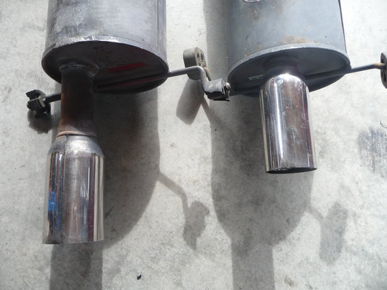 Two exhaust mufflers, one with a chrome tip, are mounted on a concrete surface