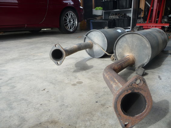 Two rusty exhaust mufflers with flanges on a concrete floor, with a red car side visible in the background