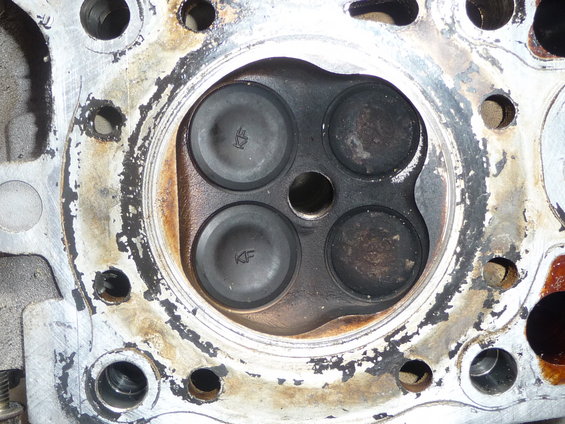 Engine cylinder head with four valves, two marked KF, showing carbon buildup