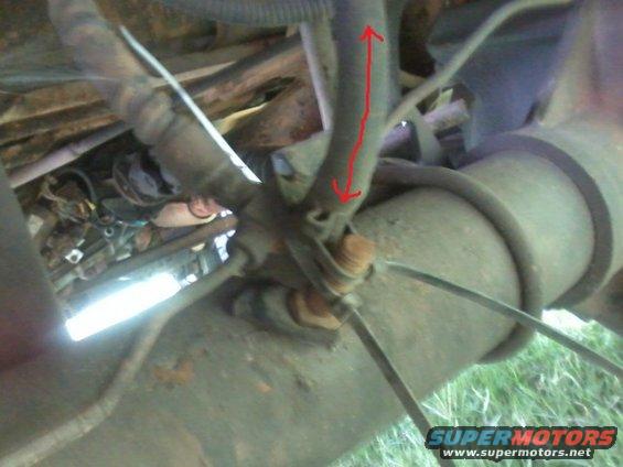Undercarriage view of a vehicle with hoses and mechanical parts; a red arrow indicates a measurement on a hose