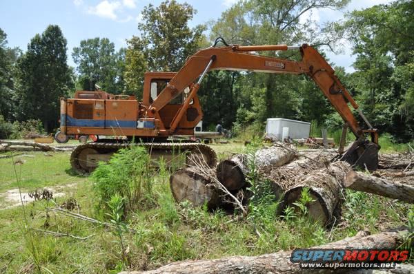 Orange excavator with tracks sits in a wooded area next to large logs