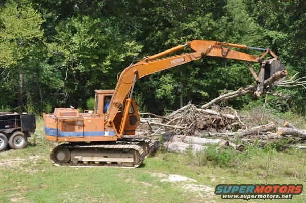 Orange excavator with blue stripe clearing logs in a wooded area