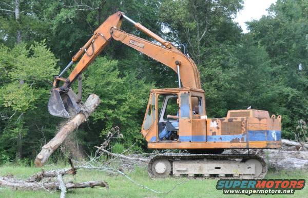 Orange excavator with blue stripe lifting a log in a wooded area