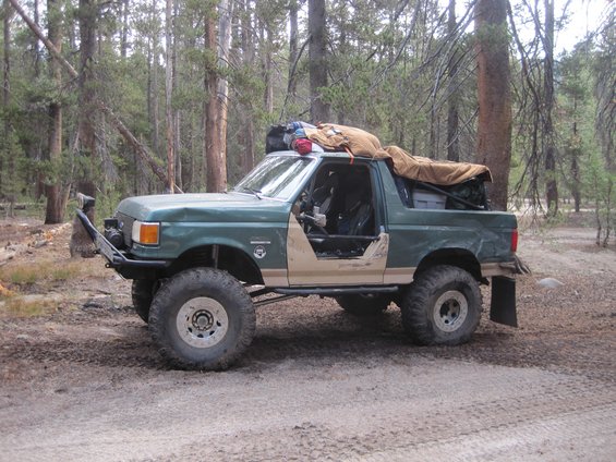 Green Ford Bronco with off-road modifications, parked in a forest. Doors removed, large tires, and gear on the roof