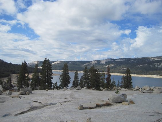 View of a calm lake surrounded by pine trees and mountains under a cloudy sky