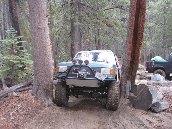Green Ford Bronco on a dirt trail in the woods, with a black truck behind it. License plate reads 2REU146