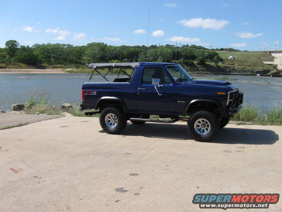 1982 Ford Bronco with a soft top frame, parked on concrete near a river and dam