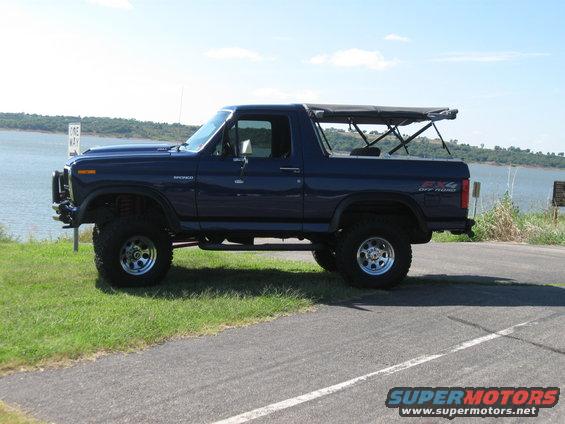 Dark blue Ford Bronco with a soft top, lifted suspension, and off-road tires parked on a roadside next to a body of water