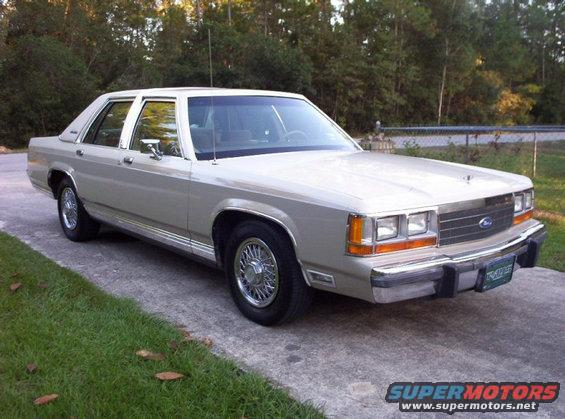 1980s Ford sedan parked on a driveway, front quarter view. Chrome trim and wire wheels are visible