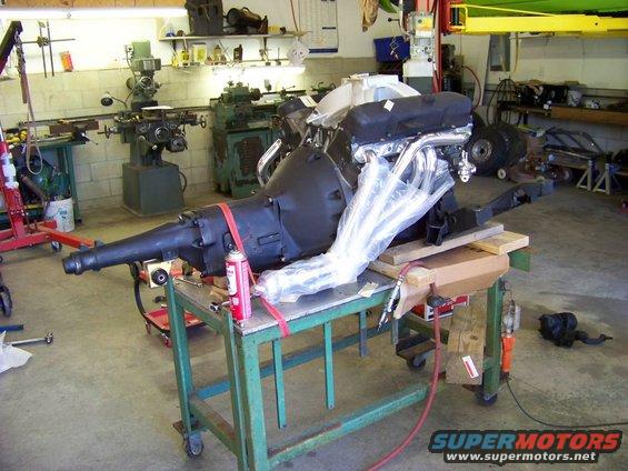 Engine and transmission assembly on a cart in a workshop. Exhaust headers are covered in plastic