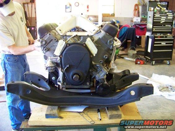 Engine and subframe assembly on wooden blocks, with a person in jeans and a t-shirt nearby
