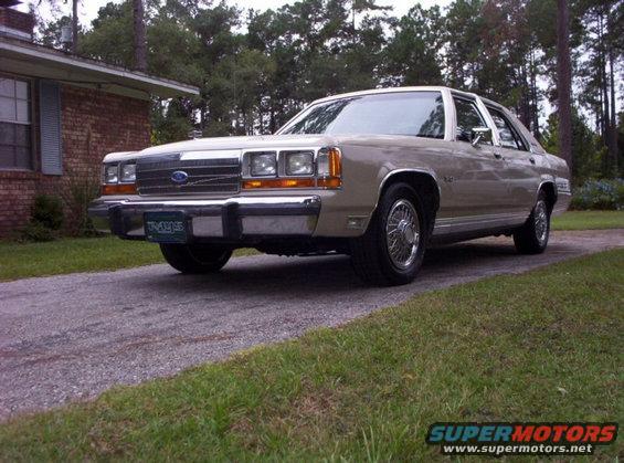 Tan 1990 Ford Crown Victoria sedan parked on a driveway, front 3/4 view. Chrome grille, chrome wheels, and a license plate are visible