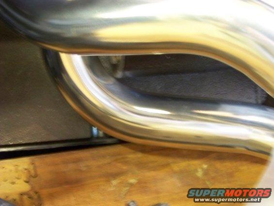 Close-up of shiny metal exhaust headers with a dark background and wooden surface below. Supermotors.net logo visible