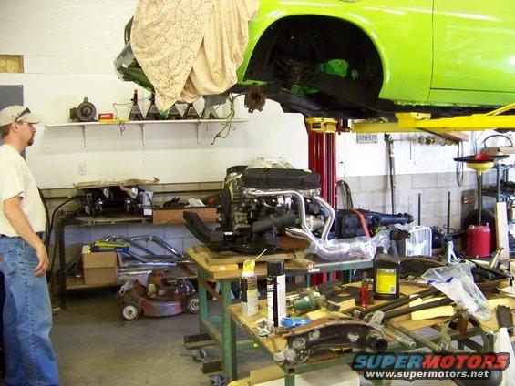 Engine on a stand with a green car body lifted above it in a garage. Parts and tools are scattered around