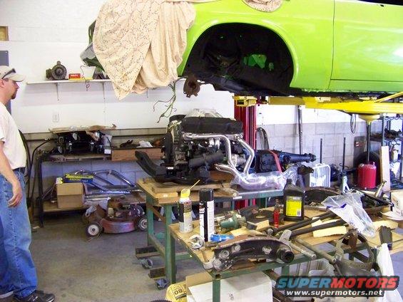 Green car body on lift, engine on table below, man watching