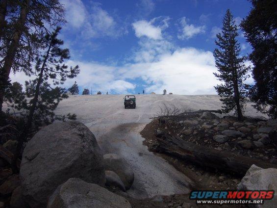 Jeep-like vehicle driving up a snow-covered slope with trees and rocks around