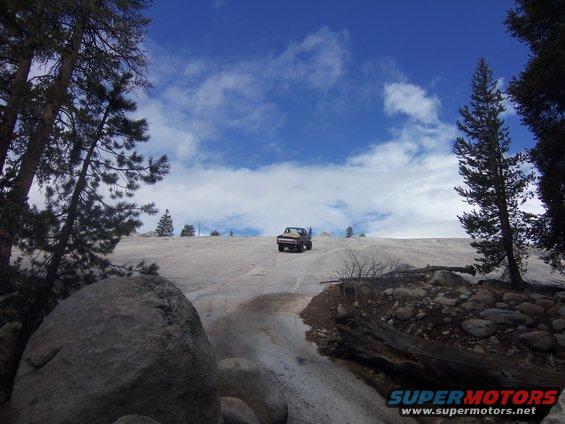 Red pickup truck driving up a snow-covered hill with trees and rocks