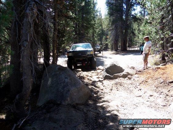 Two lifted trucks navigate a rocky, wooded trail. One truck is in the foreground, the other further back
