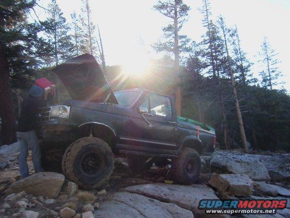 Guy working on engine of lifted Ford F-Series truck on rocky terrain with sun flare