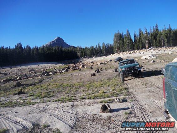 Green off-road truck driving on dirt path with trees and mountain in background. Another truck is behind it