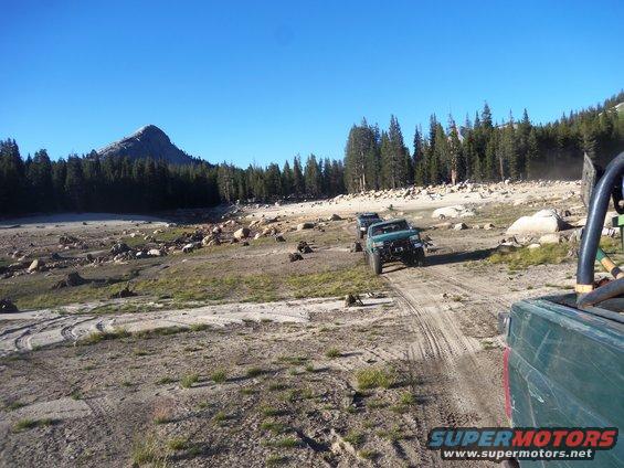 Green off-road vehicles on a dirt track with trees and a mountain in the background