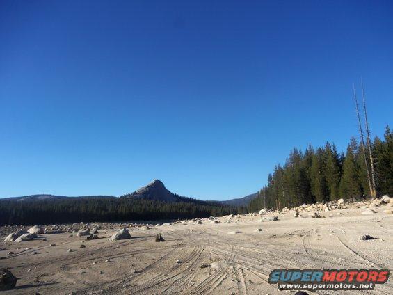Dry, rocky terrain with tire tracks leading towards a mountain and forest under a clear blue sky. Supermotors logo in corner