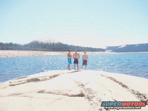 Three young men stand on a rocky outcrop by a large blue lake with trees and a mountain in the background