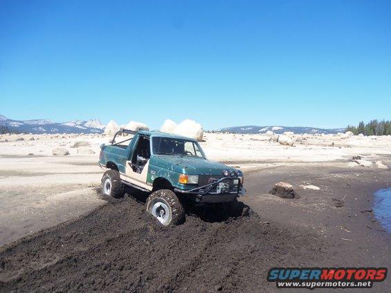 Green off-road truck stuck in mud near water, with rocky, mountainous background