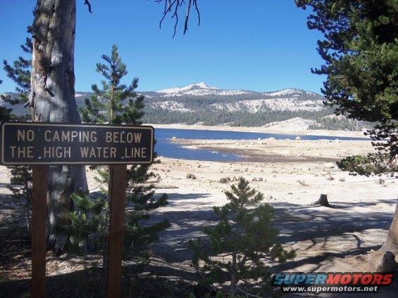 Sign reads 'NO CAMPING BELOW THE HIGH WATER LINE' near a lake and mountains