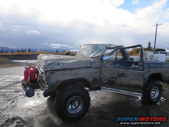Mud-covered dark-colored Ford F-Series truck with a dented hood and cracked windshield, equipped with a red jerrycan and a winch