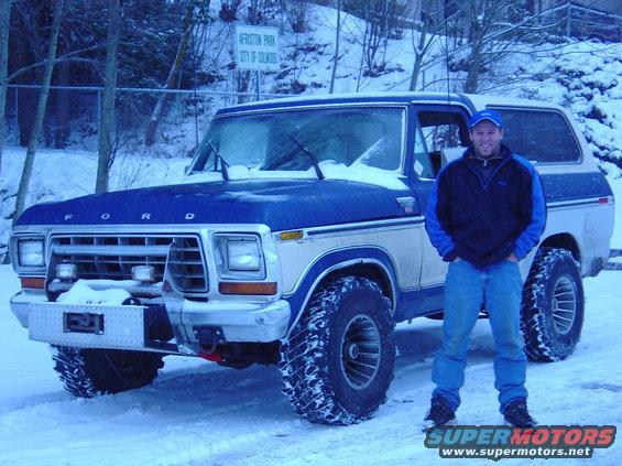 Man standing next to a blue and white Ford Bronco with large tires in the snow
