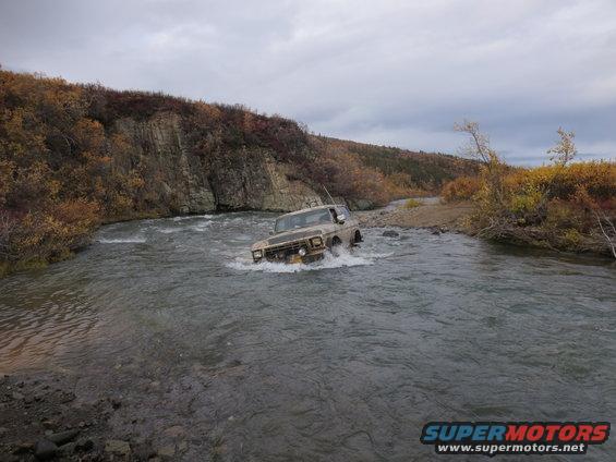 Ford F-Series truck driving through a river with rocky banks and autumn foliage