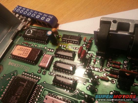 Close-up of a green circuit board with various chips, resistors, and capacitors. A set of screwdriver bits is visible at the top