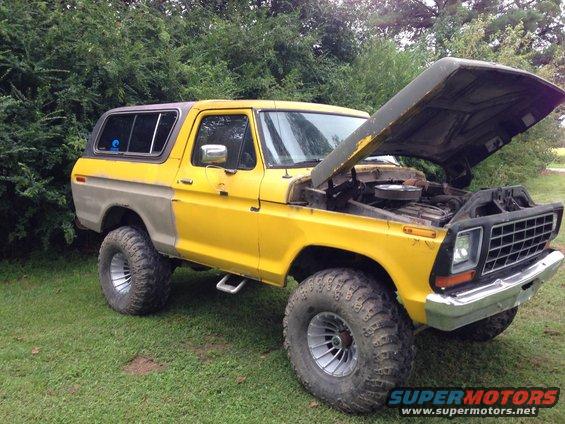 Yellow Ford Bronco with hood open, showing the engine. It has large off-road tires and a two-tone paint job