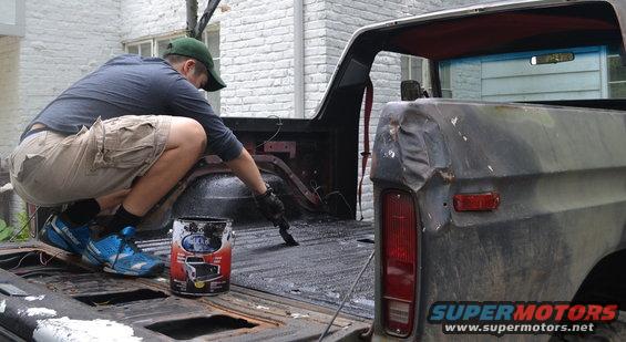 Man crouches in bed of 1979 Ford Bronco, painting the surface with a brush. Paint can visible