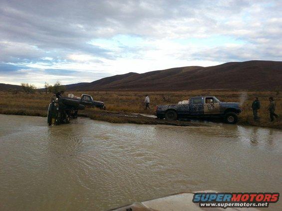 Two trucks stuck in muddy water, one on its side. Another truck is partially submerged
