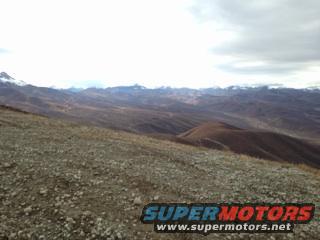 Rocky terrain with mountains in the distance under a cloudy sky. Supermotors.net logo visible