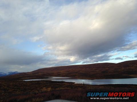 Landscape scene with a lake and rolling hills under a cloudy sky. A portion of a vehicle is visible at the bottom