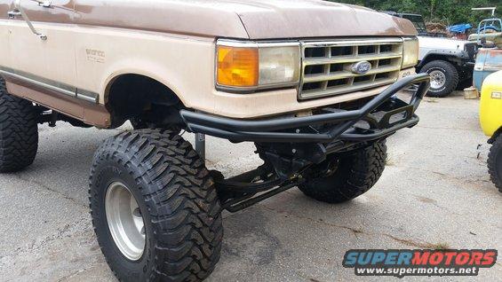 Brown Ford Bronco with big tires and a black aftermarket bumper. Another vehicle is visible to the right