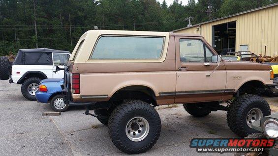 Lifted two-tone brown Ford Bronco parked next to a white Bronco and a small blue car