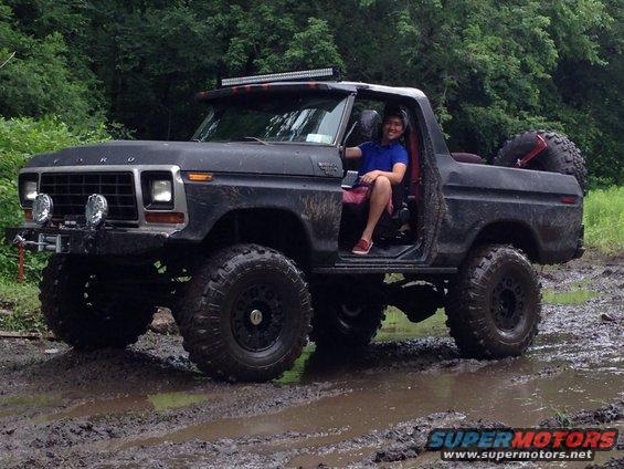 Modified black Ford Bronco with lifted suspension and off-road tires driving through mud. Driver sits in open door