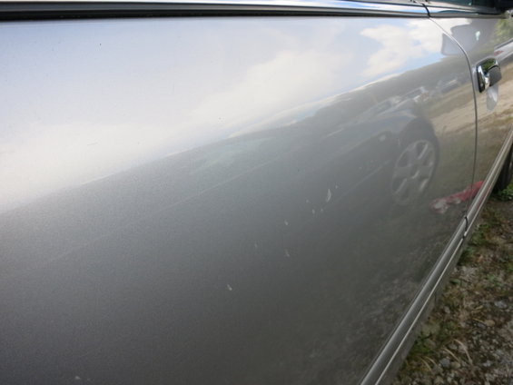 Silver car side door with scratches and reflections of a wheel and sky
