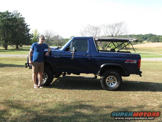 1982 Ford Bronco, blue, with a man standing next to it. It has a soft top over the rear