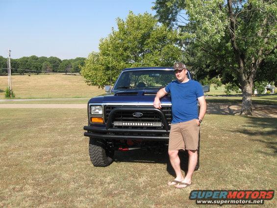 Man leaning on blue Ford truck with aftermarket bumper and light bar. Truck has square headlights and body lines consistent with early 1980s Ford