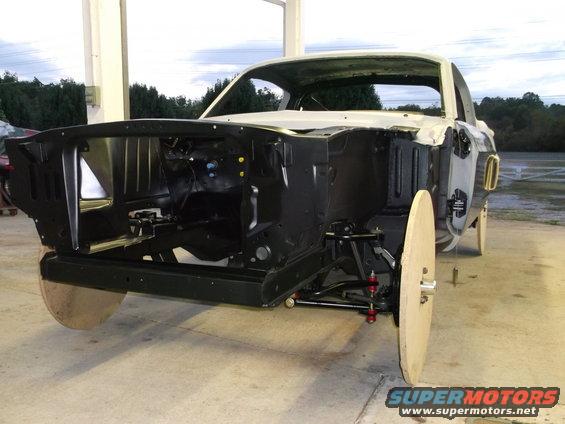 1967 Ford Mustang body in white, front view, undergoing restoration with exposed engine bay and wooden wheel covers