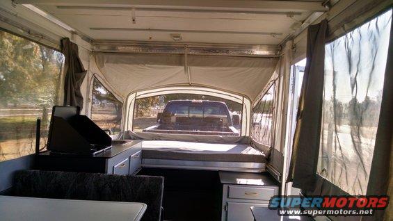 Interior of a pop-up camper showing a bed, sink, and stove, with a dark pickup truck visible through the rear window