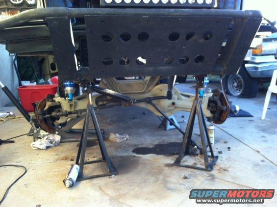 Off-road vehicle's front suspension and differential are raised on jack stands for work. Another vehicle is visible in the background